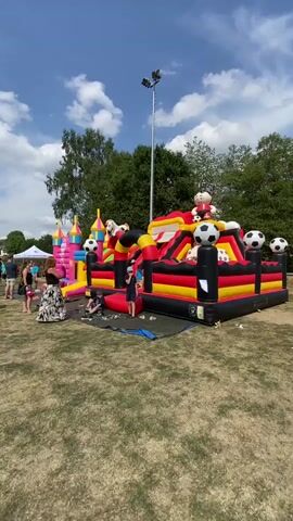 The video shows an outdoor inflatable playground with a large bouncy castle designed with black, yellow, and red colors, resembling the German flag, and decorated with soccer balls. To the left of the bouncy castle, there's a pink inflatable castle. Several people are present in the background, standing and observing. Some children are playing on the inflatable structures. The ground is covered with dry grass, and trees are visible in the distance. The sky is blue with scattered white clouds, and a tall light pole stands out against the sky. The overall scene depicts a sunny day event, likely a festival or outdoor gathering.