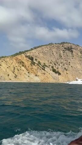 A video taken from a boat shows a large white yacht moored near a rocky, arid island. The water is choppy with white foam trailing behind the camera boat. The sky is blue with scattered clouds. The camera is positioned low, showing the edge of the boat and a person's legs wearing brown pants. The island has sparse vegetation and appears to be a cliff face.