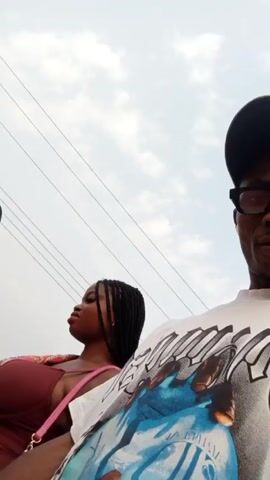 A person wearing a white t-shirt, black-rimmed glasses, and a dark-colored baseball cap is recording a video. The camera appears to be hand-held, capturing a group of three other people walking. Two women are in the foreground; one wears a brown dress and the other a maroon top and light blue jeans.  The third person, a man, wears a brown shirt and black pants. The group walks on a dirt or gravel path beside a building with a low-pitched roof. Overhead power lines are also visible.