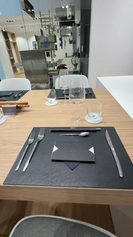 The video showcases a modern, well-lit interior, likely a showroom or high-end dining space, with a focus on a set dining table and a contemporary kitchen area.

The scene opens with a close-up of a wooden dining table set for multiple guests. Each place setting features a black placemat, silver cutlery (fork, knife, spoon), a clear drinking glass, and a wine glass. A black card or menu holder with a white abstract logo, possibly an 'X', is placed on the placemat. A small bowl of breadsticks is visible on the table. In the background, through a glass partition, parts of another bright, modern area are visible, with a person wearing a yellow top walking past.

The camera then pans right, revealing a long white counter or island that separates the dining area from a sleek kitchen. The kitchen features integrated wooden shelving with a few decorative items and a prominent built-in double oven with digital displays. The overall aesthetic is clean, minimalist, and modern. No specific text apart from the abstract logo and indistinct oven display text is clearly readable. The atmosphere is bright and organized.
