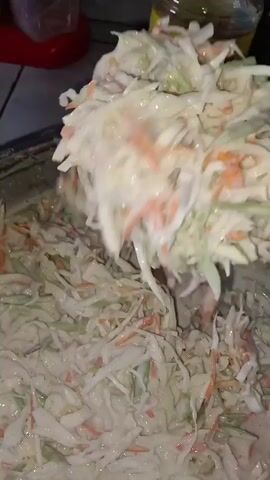The video shows a close-up shot of coleslaw being prepared in a large metal bowl. A utensil, possibly a spatula or spoon, is lifting a portion of the coleslaw, which is coated in a creamy white dressing. The coleslaw consists of shredded cabbage and carrots. The dressing is thick and appears to be dripping from the utensil back into the bowl. The background is out of focus, but a red object and a bottle of cooking oil with a yellow label can be partially seen. The action suggests the coleslaw is being mixed or served.