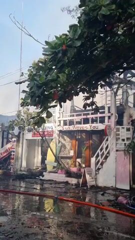 The video shows the aftermath of a fire. Several buildings appear to be significantly damaged, with some reduced to charred structures. Smoke is still visible rising from the wreckage. The ground is wet, possibly from firefighting efforts, and debris is scattered around.  Fire hoses snake across the wet surface. People are present at the scene, some walking around and observing the damage. One person is seen walking with a bicycle.  There are signs indicating businesses, but they are partially damaged and difficult to read.  Trees and other vegetation are visible in the foreground, partially obscuring the view of the damaged buildings. The sky is overcast.