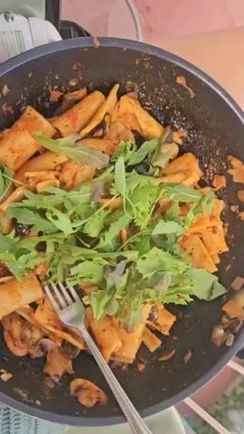 A close-up overhead shot shows a portion of pasta in a black pan, topped with green leafy vegetables and a fork. The pasta appears to be rigatoni, coated in a red sauce with visible pieces of mushrooms and possibly olives. The greens are scattered over the pasta, with some leaves showing hints of red. The pan is dark and appears to have remnants of food on its surface. A metal fork rests on top of the pasta, with its tines partially submerged. The background is slightly blurred, showing what looks like a pink surface and a glimpse of greenery. The lighting suggests an outdoor setting or a well-lit indoor space.