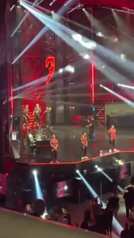 The video shows a live performance on a stage with a large crowd in attendance.  The stage is lit with bright white and red lights and features a large red numeral "2" as a backdrop. A band is visible on stage, along with several figures wearing red outfits.  During the video, pyrotechnics in the form of geysers of white smoke erupt from the stage. The crowd is dense and appears to be enjoying the show. The camera is positioned at a distance, providing a wide view of the stage and audience.