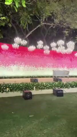 The video shows a man helping a toddler with a drink on a lawn at night. The lawn is illuminated by artificial lights, and there's a large backdrop with white and pink flowers, decorated with chandeliers. The man, dressed in a black embroidered outfit, is bending down to give the toddler a small bottle with a straw. The toddler, wearing a blue shirt, is reaching for the bottle. The man appears to be opening the bottle cap to assist the child. There are several black boxes, possibly speakers or generators, on the lawn near the floral backdrop.