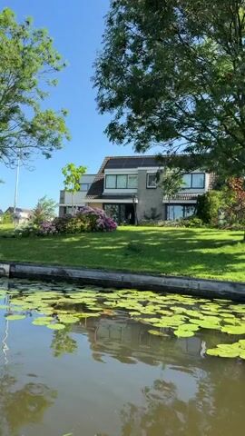 The video shows a canal with lily pads on the surface. On the bank of the canal are a grassy lawn, a modern house with large windows, and several trees. The sky is clear and blue, indicating a sunny day. The camera pans slowly from right to left, showing different parts of the scene.