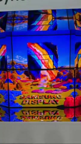 The video shows a large Samsung display made of many smaller screens showing a landscape image in vivid colors. The words "Samsung Display" are spelled out in flowers at the bottom of the image. The display is labeled "Synchroma". The camera zooms out to show the display in its entirety, surrounded by white walls. People can be seen walking in the background.