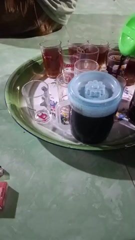 The video shows an indoor scene, likely in a home, with a light green tiled floor. A large, round, metallic green tray occupies the center of the frame, holding several items. On the tray, there are multiple small, clear drinking glasses; some are empty, while others contain an amber-colored liquid, likely tea. One glass has "MOTO" printed on it, and another "LEZZA". A clear jug with a light blue, flower-shaped lid contains a dark liquid, possibly coffee or strong tea.

An activity is taking place where a green plastic pitcher is being used to pour more of the amber liquid into one of the glasses on the tray. Partially visible in the background, a person is seated on the floor, suggesting a casual gathering. To the left of the tray on the floor, a red pack of "DIARUM" cigarettes and a green and silver lighter are visible. The lighting appears to be artificial, indicating it might be evening or nighttime. The overall scene suggests a social occasion where drinks are being served among a group of people.