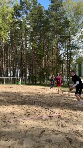 The video captures a beach volleyball game in action. Four people are visible on the sand court, divided into two teams by a net. The person in the initial frame serves the volleyball over the net, while their teammates and opponents position themselves to play. Tall trees surround the sandy court, with a blue sky and scattered clouds overhead. The court is marked with red tape, and there are structures like a ladder and a small building in the background, adding context to the outdoor setting.