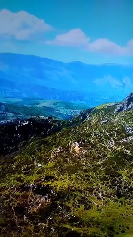 An aerial drone shot pans across a vast mountainous landscape under a bright blue sky with scattered clouds. The foreground is dominated by a rugged, green mountainside covered in low-lying shrubs and dotted with rocky outcrops. A winding dirt path is visible snaking up the slope. In the mid-ground, the mountains descend into a verdant valley dotted with small buildings and fields, suggesting a rural settlement. Further in the distance, layers of misty blue mountains fade into the horizon, creating a sense of immense depth and scale. The sunlight casts shadows, highlighting the textures of the terrain.