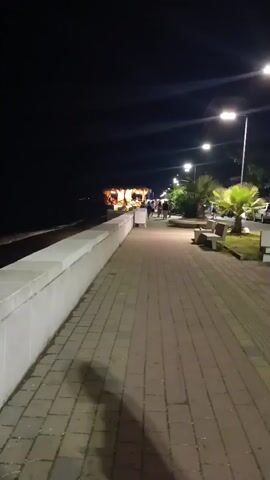 This is a first-person perspective video taken at night along a paved walkway next to a body of water. A low white wall runs along the left side of the walkway, separating it from the water. Further down the walkway, there is a thatched-roof structure with lights and people inside and around it, appearing to be a bar or restaurant. Several people are walking along the walkway, some in groups. Streetlights illuminate the path, and there are palm trees and benches visible on the right side of the walkway.