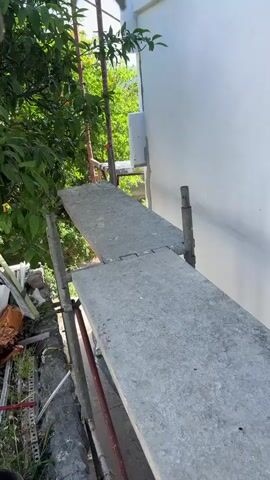 The video shows scaffolding erected against the side of a white building. On the scaffolding are concrete planks and metal poles. There is a window on the building, and it appears to be undergoing some sort of renovation or repair, with exposed insulation around it. Trees and foliage are visible on the left side of the frame, and a bright blue sky is visible at the top. In the second and third segments, a ladder is also visible. The video appears to be taken from a low angle looking upwards.