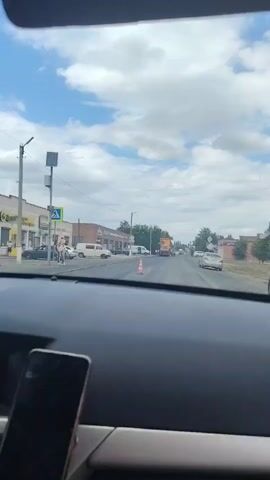 The video shows a view from inside a car looking out at a street scene. The dashboard of the car is visible in the foreground, with a phone mounted on it. Outside, there are several buildings, some with awnings, and cars parked along the side of the road. A pedestrian crossing is visible with a sign. Further down the street, there is a large orange truck with a work crew around it, appearing to be involved in road construction or repair. There are also other vehicles and trees lining the street. The sky is blue with scattered clouds.