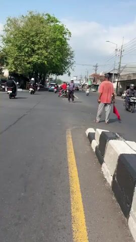 This media file captures a street scene in Siring, East Java. The video shows a road with several motorbikes and a few pedestrians. A large tree with lush green foliage dominates the center of the frame, casting a shadow on the road. The sky is blue with some scattered clouds, suggesting fair weather.

The main activity appears to be traffic on a local road. Several people are riding motorbikes, some of whom are wearing helmets. One person in a red shirt is standing in the middle of the road, seemingly directing traffic or waiting for something. Another person in a pink shirt and gray pants is walking on the side of the road. Buildings line the street, indicating a populated area. The time of day appears to be daytime, likely late morning or early afternoon, given the bright sunlight and shadows.

There is no visible text in the media file. The overall mood seems to be a typical, everyday scene of street life in a town in East Java. The focus is on the mundane movement of people and vehicles in a bright, outdoor environment.