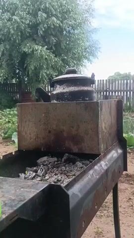 A kettle sits on a makeshift metal enclosure atop a barbecue grill. The grill contains burning coals and ash. The background shows trees and a wooden fence. The video is static.