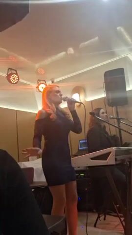 A woman in a dark blue dress sings into a microphone on a small stage, illuminated by rotating green, blue, and purple stage lights. She is blonde and stands to the left of a woman sitting and playing a white keyboard. A black speaker is behind the keyboard. The venue has light wood paneling and a white ceiling with embedded lighting. Tables with white napkins are visible to the left, suggesting a restaurant or event space setting. A blurry, dark figure of an audience member is partially visible in the foreground.
