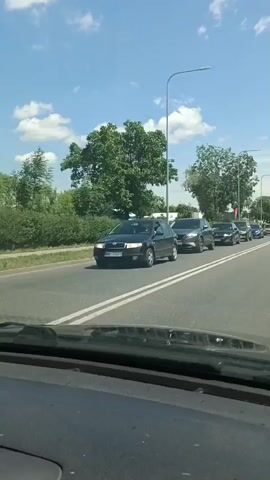 The video shows a view from inside a vehicle traveling on a road. The road is asphalt with white lane markers. On the right side of the road, there is a line of cars. The first car visible is a dark-colored SUV. Following it is a dark-colored sedan, then a blue sedan, and then other vehicles further down the road. On the left side of the road, there is a dark-colored car passing by in the opposite direction. The road is lined with trees and greenery. The sky is blue with scattered white clouds. There are streetlights visible along the side of the road. The video appears to have been recorded during the daytime.
