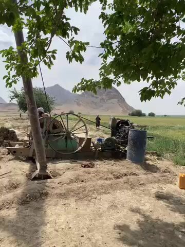 The video captures a well-drilling operation in an arid, open landscape, likely in Nowruzi, Afghanistan. In the foreground, a large drilling rig with a prominent green wheel and an engine is actively running, partially shaded by a leafy tree. Several long pipes are laid out on the dusty ground. One man, dressed in dark clothing, walks towards these pipes from the left side of the frame. Another man is visible closer to the drilling rig, and a third figure can be seen in the distance near the green vegetation. A blue barrel sits beside the machinery. The setting features a clear sky with scattered clouds, and towering mountains form the backdrop in the distance. The overall scene suggests a bright, sunny day. The activity is focused on the operation of the drilling rig, with pipes being handled for the well.