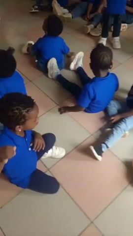 The video shows a group of young children, likely preschoolers or kindergarteners, gathered indoors. Most of the children are sitting on a tiled floor, while some stand and move around. They are predominantly wearing blue polo shirts, which appear to be a uniform, paired with jeans or trousers. Some children have white sneakers, others dark shoes, and one child is wearing light brown boots.

An adult, possibly a teacher, is visible in the background, standing and facing the children. The children are engaged in an activity, clapping their hands and appearing to sing or follow instructions. Their expressions suggest they are happy and enjoying the activity. The setting appears to be a classroom or a school hall, well-lit, indicating it is likely daytime. There is a small logo visible on the blue polo shirts, but it is not discernible.