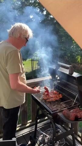 A person with blonde hair and glasses, wearing a light green t-shirt and dark pants, is standing on a wooden deck and grilling meat on a charcoal grill. Smoke billows from the grill as the person uses tongs to turn pieces of meat. A large cut of raw meat sits on a platter to the right of the grill. Trees and foliage are visible in the background, and a beige awning extends over the deck. The sun is shining, casting light on the scene.