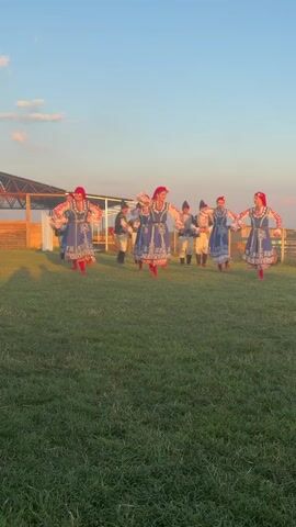 A group of eight people, dressed in traditional attire, are dancing in a grassy field. The individuals in the foreground are women wearing blue dresses with intricate white and red embroidery, red headscarves, and red shoes. The individuals in the background appear to be men wearing white shirts, dark vests, and dark hats. The group is performing a synchronized dance routine. The sun is setting, casting long shadows across the grass. In the background, there is a wooden fence and a covered structure. The sky is a clear blue with a few wispy clouds.