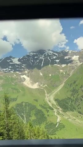 The video shows a scenic view of a mountain range covered in snow on its peaks. Lush green slopes descend from the mountains, with a winding path visible on one of them. Tall, dark green coniferous trees are in the foreground, partially obscuring the view of the mountains. The sky is blue with scattered white clouds. The camera appears to be moving, likely from a vehicle, as indicated by the slight blur and the framing of the scene through what seems to be a window. The overall impression is of a beautiful, natural landscape.