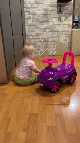 The video shows a toddler walking around a room. The room has a grey brick wall and a wooden floor. There is a purple toy car on the floor near a grey cabinet. A blonde toddler wearing a pink Minnie Mouse t-shirt and green pants is initially bent over the car, then stands up and walks away from it.