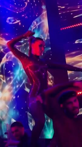 A woman in a sparkling dark leotard with cutouts dances on a stage under red and blue lights. A projection of colorful, abstract shapes is behind her. Two men, one in the foreground to the lower right, and another to the lower left, are visible watching the dancer.