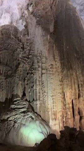 The video pans across a large cave formation with numerous stalactites and stalagmites. The formations are various shades of brown and white, with some areas illuminated by artificial lights, casting yellow and green hues on the rocks. The camera moves slowly, revealing the scale and complexity of the cave's natural structures. The environment appears to be a tourist attraction, with pathways and lighting suggesting a managed experience. No people are visible in the video.