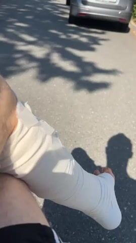 A close-up shot shows a person's leg wrapped in a white cast, resting on the asphalt. The camera slowly pans up and to the right, revealing a street lined with trees and buildings. Two cars are parked on the right side of the street. Sunlight filters through the leaves, casting shadows on the ground. The scene appears to be a bright, sunny day in a residential or urban area.