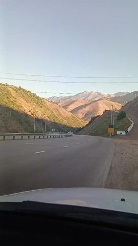 The video captures a forward-facing view from inside a vehicle as it travels along a paved road that curves to the right. The road is flanked by guardrails on the left and a dirt shoulder on the right. Beyond the guardrails, the landscape transitions into a steep, vegetated hillside. In the distance, majestic mountains with exposed rock faces dominate the horizon under a clear sky. The sun is low, casting long shadows and illuminating the scene with warm light. A few other vehicles are visible on the road ahead, traveling in the same direction. There are also utility poles and lines visible along the roadside. Traffic signs are present on the right side of the road. The overall impression is a scenic drive through a mountainous region.