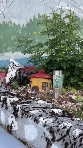The video shows a close-up view of a garden decoration with a mural in the background. In the foreground, there is a stone wall with some moss and white paint splatters. Behind the wall, there are small decorative houses, one yellow with a red roof and a window, and another partially visible. There are also two solar-powered garden lights with clear textured covers and silver bases. Lush green plants and small pink and yellow flowers are growing around the decorations. The background mural depicts a green landscape with a blue body of water and mountains. A black bird silhouette is visible on the mural. The scene appears to be outdoors in a garden or yard setting.