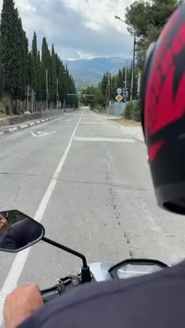 The video shows a point-of-view shot from a motorcycle, moving along a paved road. The road is lined with tall, manicured cypress trees on the left side, with a metal fence separating the trees from the road. On the right, there are more trees and a metal fence. In the distance, mountains are visible under a cloudy sky. The sky is bright, suggesting daytime.

The road has white and yellow pedestrian crossings. Traffic signs are visible, including a "STOP" sign and a traffic light. The rider is wearing a red and black helmet. The overall scene appears to be a scenic route, possibly in a rural or semi-rural area, given the presence of mountains and natural landscaping. The activity is simply riding a motorcycle down the road. No specific event or interaction is depicted, just the journey itself. The mood is calm and observant, as one might feel while taking in a scenic drive.
