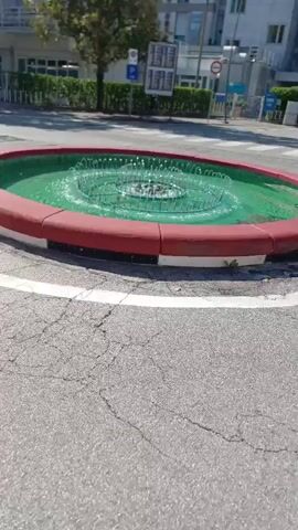 The video shows a circular water fountain in the middle of a roundabout. The fountain has a red border and is filled with green water. The water is being sprayed upwards from a central mechanism, creating ripples and waves. The roundabout is paved with asphalt and has white markings on the road. In the background, there are buildings and trees. The camera is tilted at an angle, making the scene appear dynamic.