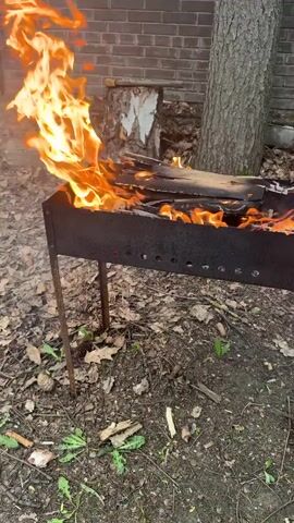 A metal grill is ablaze with a lively fire. The flames are bright orange and yellow, leaping upwards from the burning wood inside the grill. The grill itself is black and has a series of holes along its sides. It stands on four thin metal legs on a ground covered with fallen leaves and patches of green vegetation. Behind the grill, there is a brick wall and the trunk of a tree. A cut tree stump is visible to the left of the tree. Smoke rises from the burning wood.