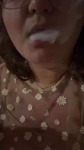 A close-up video shows a person exhaling a cloud of white smoke. The person is wearing glasses and a sheer shirt with a daisy pattern. The smoke obscures part of their face and chest.