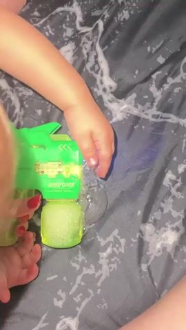 A child's hand holds a green bubble gun. Bubbles are being released from the gun and floating in the air. The background is a dark, patterned fabric. The lighting is dim.
