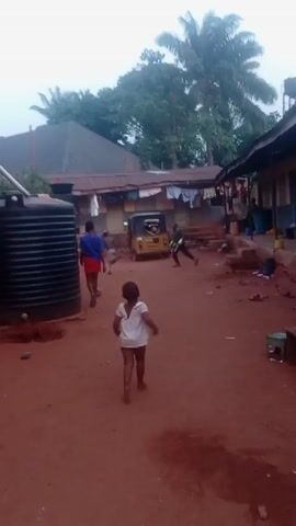 The video shows a red dirt courtyard surrounded by buildings in what appears to be a rural area. In the courtyard, there is a large black water tank on the left. A young boy in a blue shirt and red shorts walks across the courtyard from left to right. A small girl wearing a white shirt walks in the same direction, ahead of the boy. In the background, there's a yellow three-wheeled vehicle. Clothes hang on a line strung between buildings in the background.  There are benches and other objects scattered around the courtyard. Trees are visible in the background.
