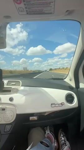 The video begins with a view from inside a car, looking out the windshield at a highway. The sky is blue with scattered white clouds. The dashboard of the car is visible in the foreground, with a white finish and a "500" logo. The driver's feet are visible, wearing sneakers. The camera then pans to the side to show a woman with blonde hair, wearing sunglasses and a pink shirt, smiling and taking a selfie. She is in the passenger seat of the car, with a seatbelt on. The car appears to be moving on a highway.