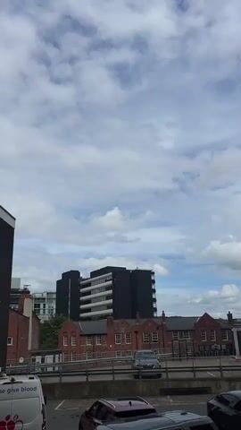 The camera pans up from a parking lot, revealing a view of several buildings. The parking lot contains various cars, including a van with "give blood" written on it. In the background, there are two main buildings: a taller building with a grey facade and multiple windows, and a shorter building that is brick and red in color. The sky is visible, filled with clouds, some blue patches, and a generally overcast appearance. The tall building is rectangular and appears to be an office building. The video is taken from a low angle, emphasizing the height of the buildings.