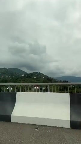 The video shows a view from a vehicle driving on a road. In the foreground, a concrete barrier with black and white stripes is visible. Beyond the barrier, a metal railing runs horizontally across the frame. In the background, a lush green mountainside dotted with houses and trees stretches out under a cloudy sky. The clouds are dark and appear heavy, suggesting an overcast or possibly rainy day. The overall impression is of a scenic, rural landscape viewed from an elevated position, likely a bridge or overpass.
