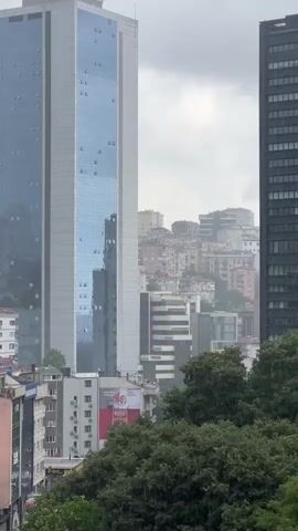 A video shows a cityscape with modern buildings and older, densely packed residential buildings in the background. Two tall buildings, one white with blue glass and another dark gray, dominate the foreground. Green trees are visible in the lower part of the frame, obscuring some of the lower buildings. The sky is overcast and appears to be raining, with visible streaks of rain in the air. Several cars are parked on a sloped area on the right side of the frame. There are no people clearly visible in the video.