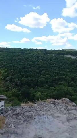 The video pans across a rocky landscape with a lush green valley below. The sky is blue with scattered white clouds. The camera then moves to show ancient stone structures carved into the cliff face, some with visible cave entrances. The path ahead is paved with rough stones, winding uphill through vegetation. The video captures a natural and historical environment.