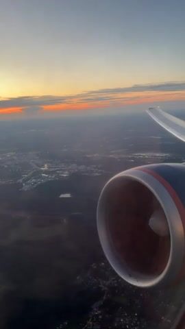 This is a vertical, first-person perspective video shot from the window of an airplane during what appears to be sunset. The engine of the plane is visible in the foreground on the right side of the frame, a large cylindrical object with a red and orange rim. Beyond the engine, the wing of the plane extends outwards towards the right. The sky above is a gradient of pale blue and soft yellow, with a thin band of orange and red along the horizon, indicating the setting sun. Below the plane, a sprawling landscape unfolds, featuring a mixture of urban development with illuminated buildings and roads, and darker areas of trees and vegetation. A body of water, possibly a lake or river, is visible in the mid-ground. The overall impression is one of flight over a populated area as daylight fades. There are no people visible in the video, and no audio is present.