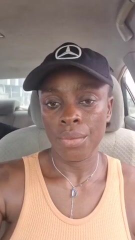 A woman with dark skin, wearing an orange tank top and a black baseball cap with a white Mercedes-Benz logo, is sitting in the driver's seat of a Toyota Camry. She has a silver necklace with a "D" pendant. Her nails are painted orange. She is driving on a city street with other vehicles visible in the background. The car's interior is visible, including the steering wheel, dashboard, and seats. She looks directly at the camera and then glances at the road while driving.