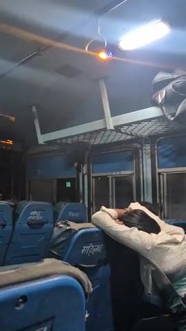 This is a video taken inside a bus, likely at night given the artificial lighting and lack of external daylight. The bus appears to be in motion, as indicated by the slight blur in some of the frames. The subjects are primarily the seats of the bus, which are blue and appear to be made of a synthetic material. Some seats are covered with dark fabric. There is a passenger visible in the foreground, leaning their head on their arm and sleeping. Their face is not clearly visible, but they are wearing a light-colored top and possibly jeans. Another passenger is visible further back in the bus, also seemingly asleep. Luggage is stowed above the seats.

The setting is clearly the interior of a public transport bus, with overhead compartments for luggage and individual seats arranged in rows. The lighting is a mix of cool white overhead lights and some warmer, colored lights, possibly from decorative elements or external sources reflecting through the windows. The windows themselves show a dark, indistinct exterior, suggesting it's nighttime or a very dimly lit area.

There is text visible on the seats, written in what appears to be Hindi. Some legible words include "मोहिनी" (Mohini) and possibly "सनातनी" (Sanatani) or similar religious/sectarian terms. Above the windshield, there is a digital display showing text in Hindi. In one frame, this display reads "जेनक दरों से सावधान" (Janak daron se savadhan), which translates to "Beware of Janak rates" or "Beware of public rates." Another frame shows "से सावधान" (se savadhan), meaning "beware of." The context of these messages is unclear without further information, but they could be warnings or advertisements.

The overall mood conveyed is one of travel and rest, with the sleeping passenger suggesting a journey that has been ongoing for some time. The presence of luggage indicates that people are traveling. The location context of Kullu, India, suggests this bus is part of the public transport network in that region, possibly a long-distance or intercity route.