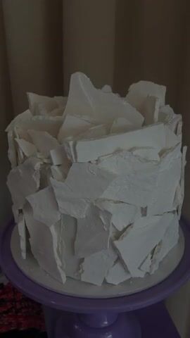 A cake decorated with white shards of what appears to be hardened icing sits on a purple cake stand. The cake has a mostly round shape and is covered with the white shards, giving it a textured and somewhat abstract look. The background is out of focus and appears to be a curtain, suggesting an indoor setting.