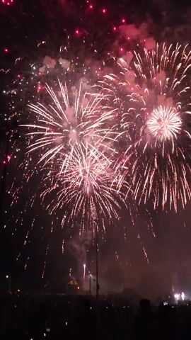 The video captures a fireworks display against a dark night sky. Several large, bright fireworks are exploding, creating bursts of white and gold light with trailing sparks. The fireworks are varied in size and intensity, with some appearing as full, rounded bursts and others as more elongated cascades. Smoke from the explosions drifts through the sky, obscuring some of the detail. Below the fireworks, a silhouetted crowd of people is visible, suggesting an outdoor event or celebration. Streetlights or other ambient lights can be seen in the distance, faintly illuminating the lower portion of the scene. The overall impression is one of a vibrant and energetic spectacle.