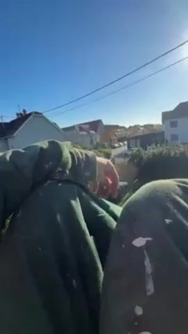 The video shows a person sitting down on what appears to be a roof or elevated position, overlooking a residential neighborhood on a bright, sunny day. The person is wearing green clothing with noticeable paint splatters. In the background, there are houses, trees, and power lines against a clear blue sky. The sun is shining brightly, with a lens flare visible.