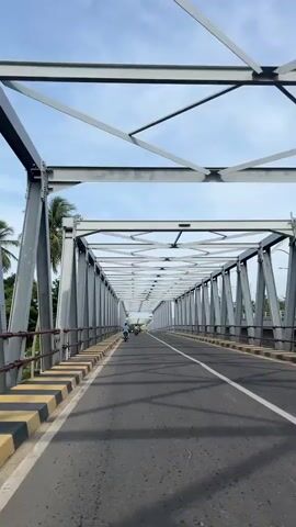 A low-angle, first-person perspective video shows an empty road stretching across a long, metal truss bridge. The sky is blue with scattered white clouds. The bridge has a grey metal structure with diagonal supports and horizontal beams forming a geometric pattern overhead. On the sides of the road are concrete barriers painted with alternating black and yellow stripes. Several people on motorcycles are visible in the distance, heading towards the end of the bridge. Palm trees and green foliage are visible on the left side of the bridge. The road is paved and has a white line down the center. Shadows from the bridge structure are cast onto the road.